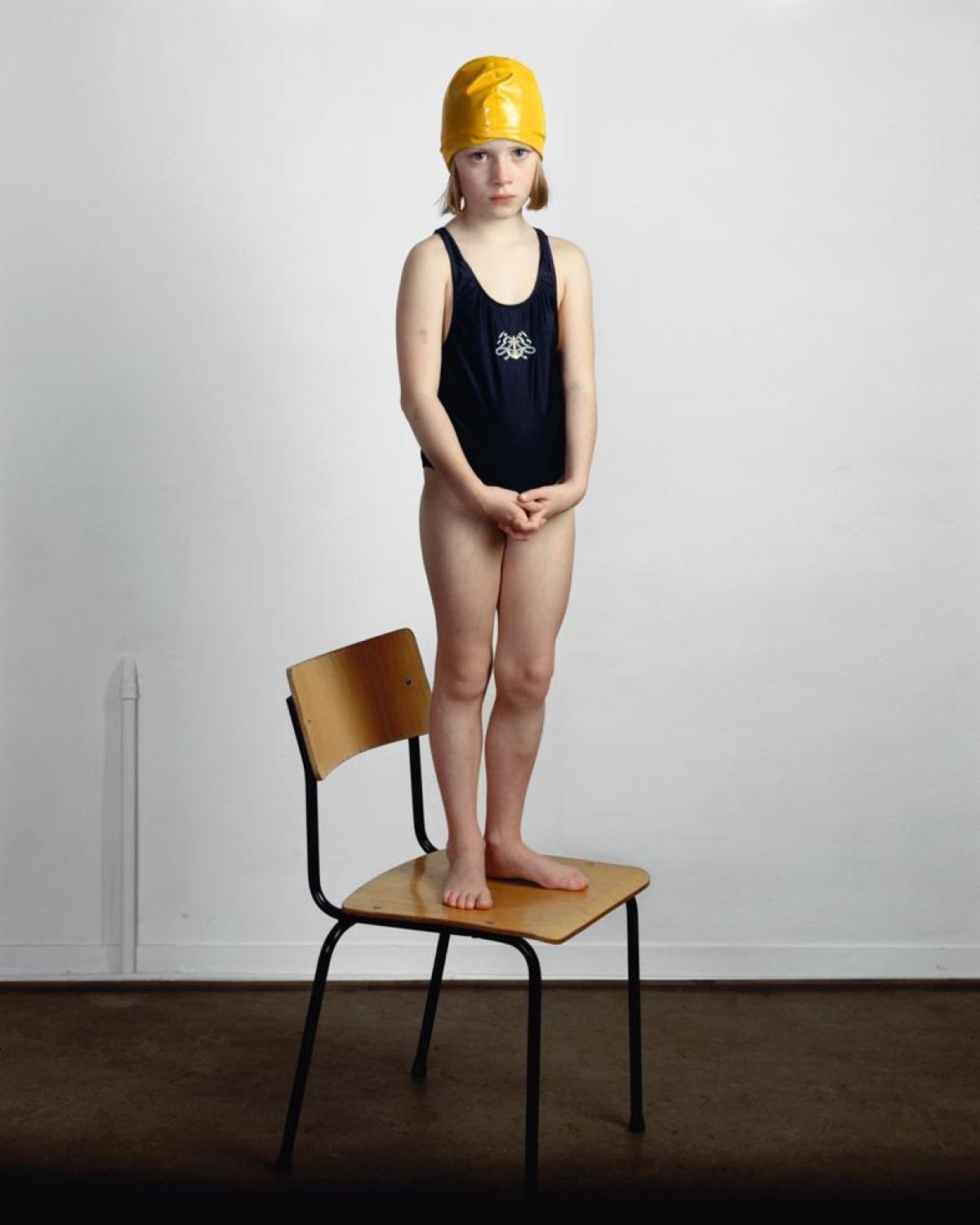 A young child in a black swimsuit and bright yellow swimming cap stands barefoot on a wooden chair with black metal legs, positioned against a white wall. The child's hands are clasped in front, gazing directly at the camera in a contemplative pose.