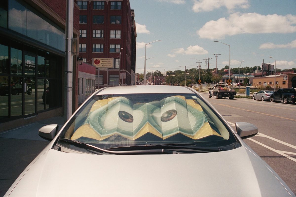 A white car parked on an urban street displays a sun shade featuring a stylized illustration of large eyes with yellow and green coloring, creating an anthropomorphic presence on the vehicle's windshield. Brick buildings, storefronts, and a clear sky line the street behind it.