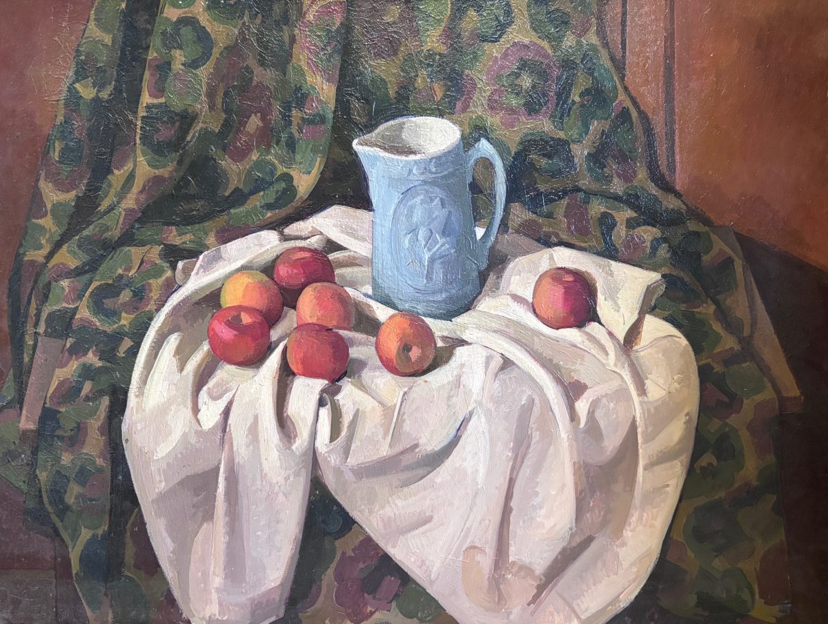 A still life composition featuring red apples arranged on white draped fabric, with a blue-grey pitcher positioned behind them. The arrangement sits against a richly patterned backdrop of dark fabric with multicolored floral motifs, set within a warm reddish-brown interior space.