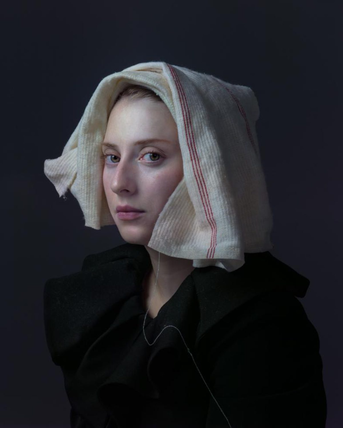 Portrait of a young woman against a dark background, wearing a black garment and a cream-colored linen head covering with a red stripe. Her gaze is directed slightly away from the camera, with a composed, contemplative expression.