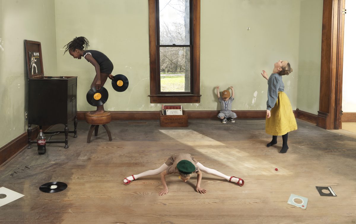 Four figures arranged throughout a pale green-walled room engage with vinyl records as sculptural and performative objects. A person in a black shirt balances on a wooden stool while holding records, another lies horizontally on the floor in a split stretch wearing a green beret, a small child sits in the center, and a figure in a yellow skirt stands to the right with arms raised. Vinyl records and related ephemera are scattered across the wooden floor, while a window frames bare trees outside.