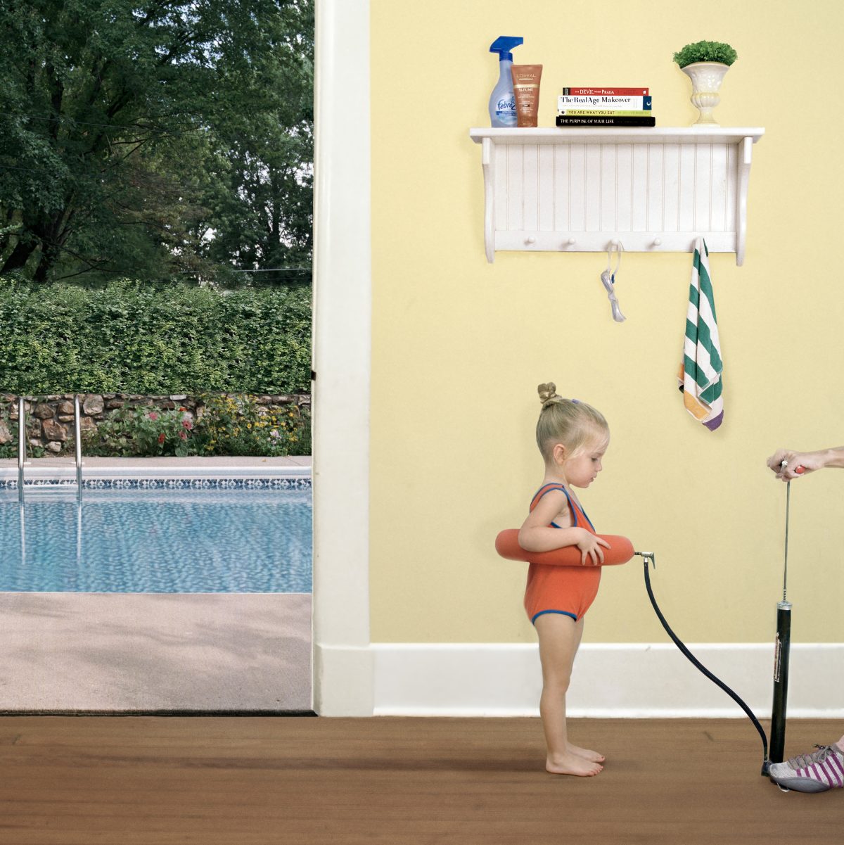 A young child wearing an orange swim floatie stands on a wooden floor facing a pale yellow wall, gazing toward an open glass door that frames a backyard swimming pool surrounded by green hedges. A white shelf mounted on the wall above holds books and household items, with towels and a hand pump hanging below, while pool maintenance equipment sits on the floor beside the child.