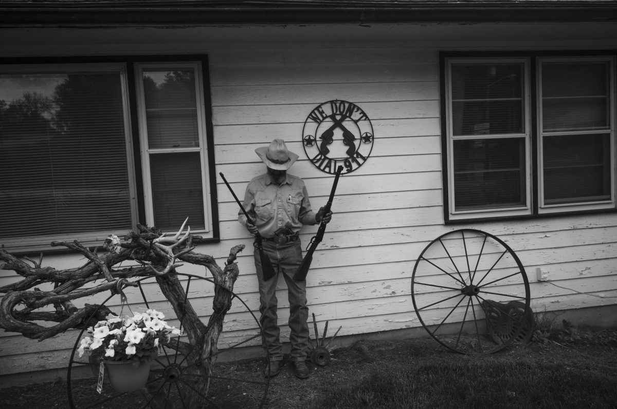 Black and white photograph of a park ranger in uniform and wide-brimmed hat standing against a wooden building wall, holding an ornate metal sign above his head. Rustic objects surround him including wagon wheels, antlers, and a potted plant with white flowers, arranged as if in a folk art or Western-themed display.