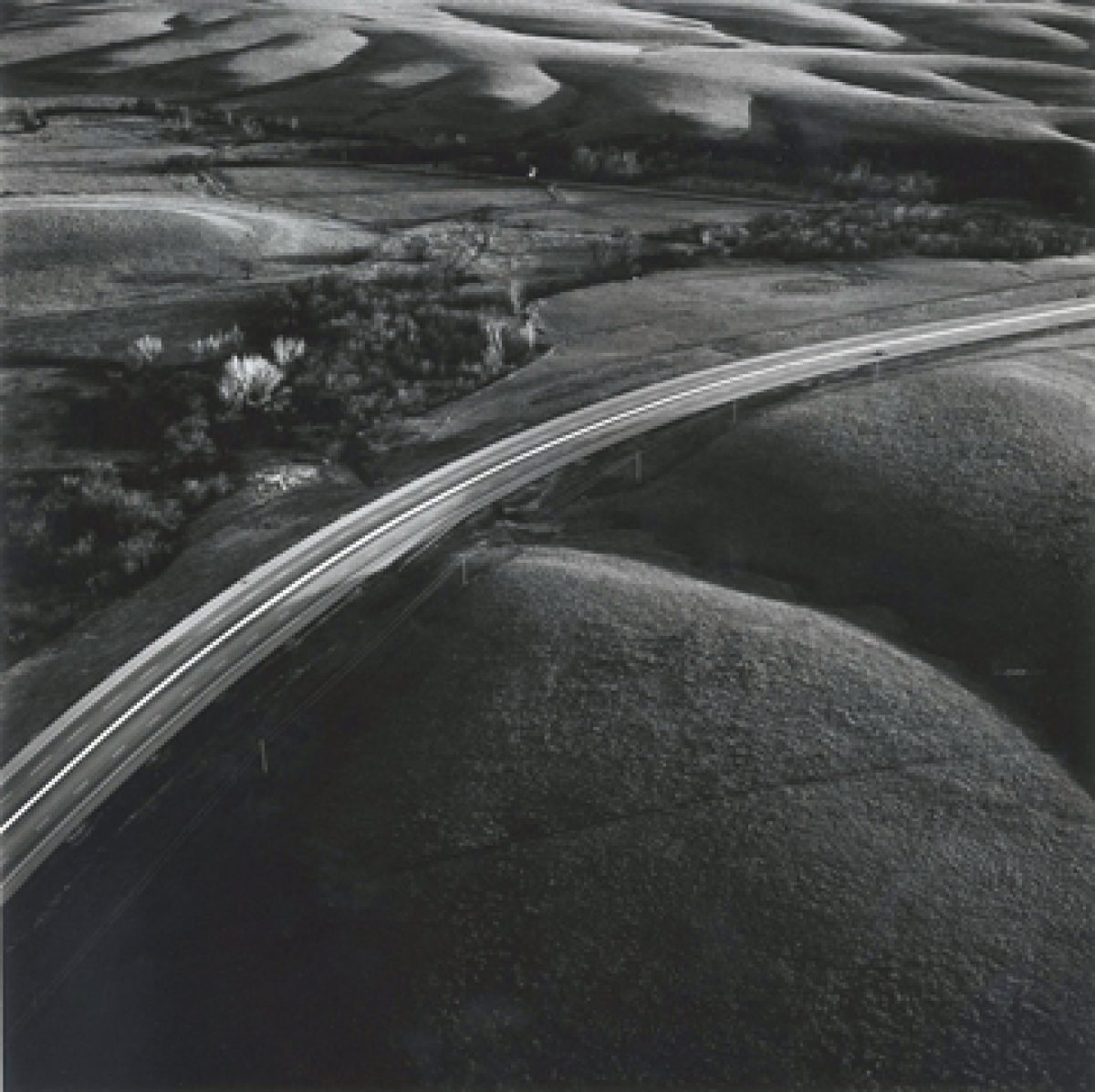 Aerial black and white photograph of a curved highway cutting through rolling prairie landscape, with agricultural fields visible in the distance and a cluster of trees and buildings nestled in a valley alongside the road.