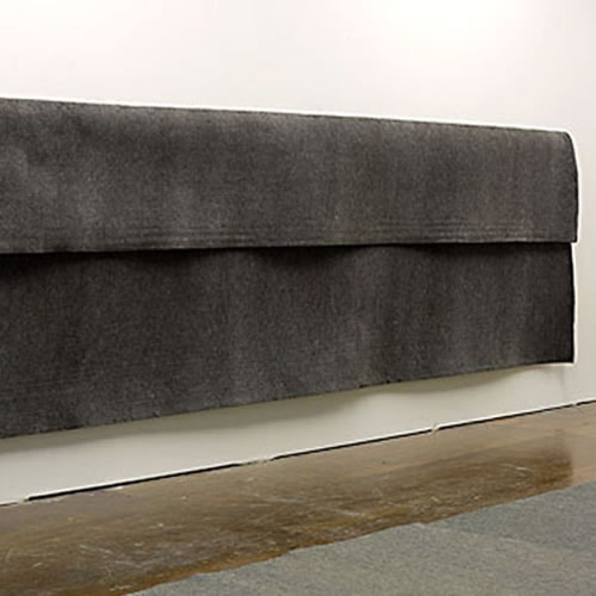 Large sheet of dark gray industrial felt suspended horizontally from a wall, with a pronounced fold creating a three-dimensional drape. The fabric hangs against a white wall above a wooden floor, emphasizing its weight and textural surface.