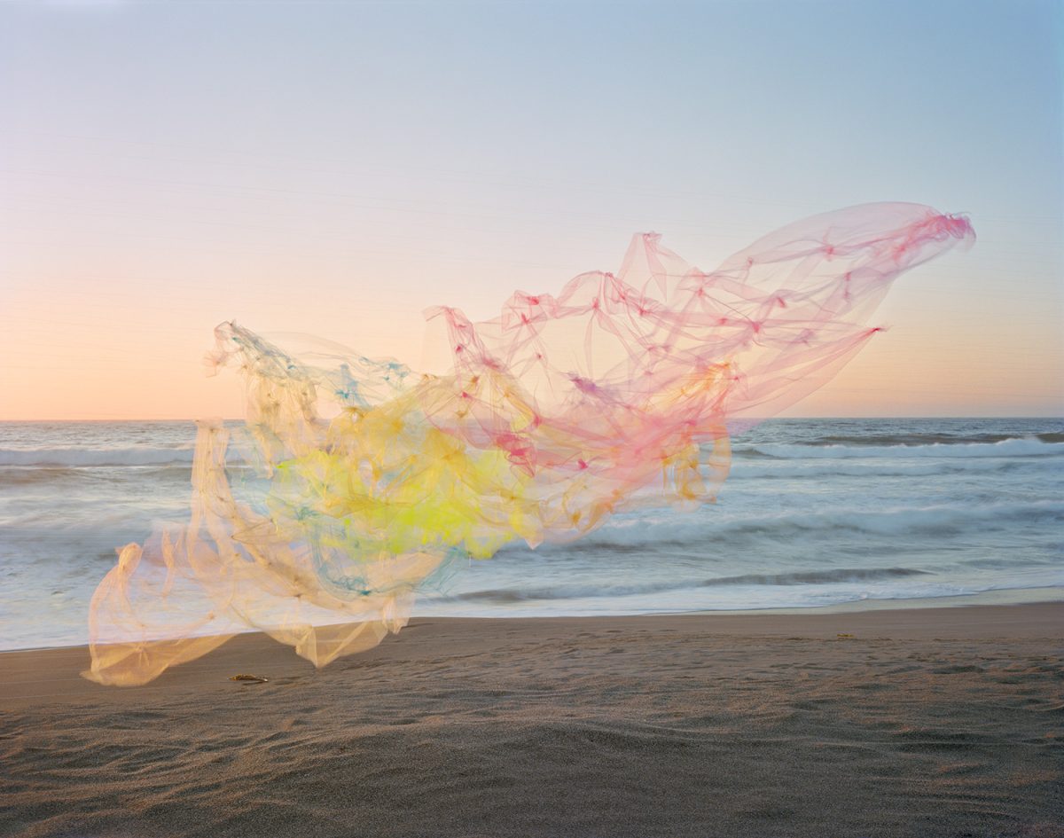 Billowing tulle fabric in soft gradations of pink, yellow, and blue floats across a beach at golden hour, with the ocean and horizon visible beyond. The translucent, flowing material creates an ethereal, sculptural form against the sand and seascape.