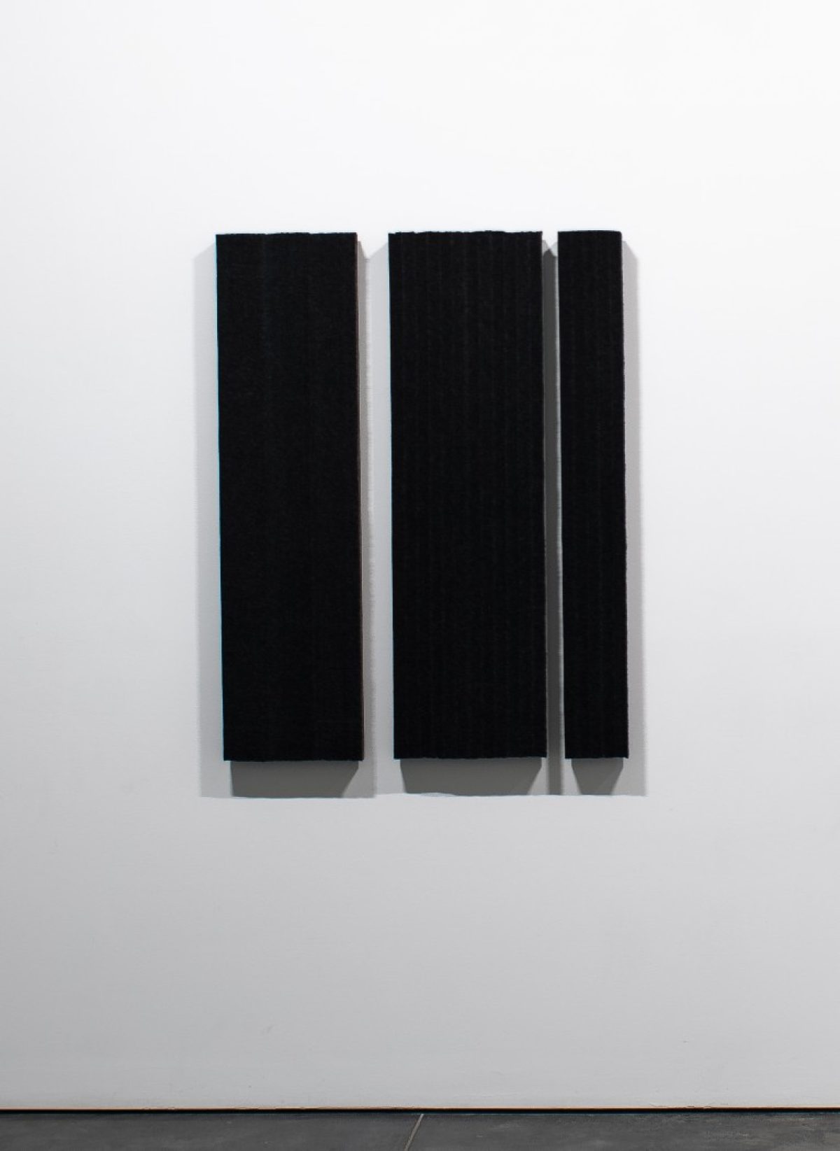 Three vertical rectangular panels of black wool felt mounted on a white gallery wall, with subtle shadows cast beneath each panel creating depth and dimension against the minimalist composition.