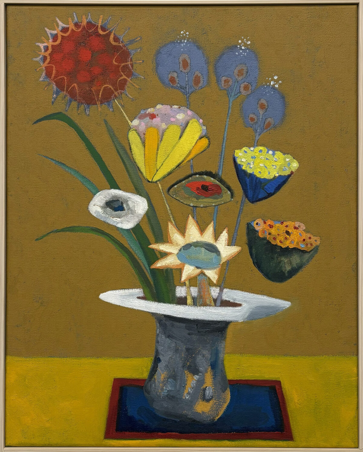 Stylized floral arrangement in a gray pedestal vase set against a mustard-gold background, featuring whimsical blooms with eye-like centers in red, yellow, white, and blue, accompanied by floating seed pods and botanical forms rendered in bold, saturated colors and primitive figurative style.