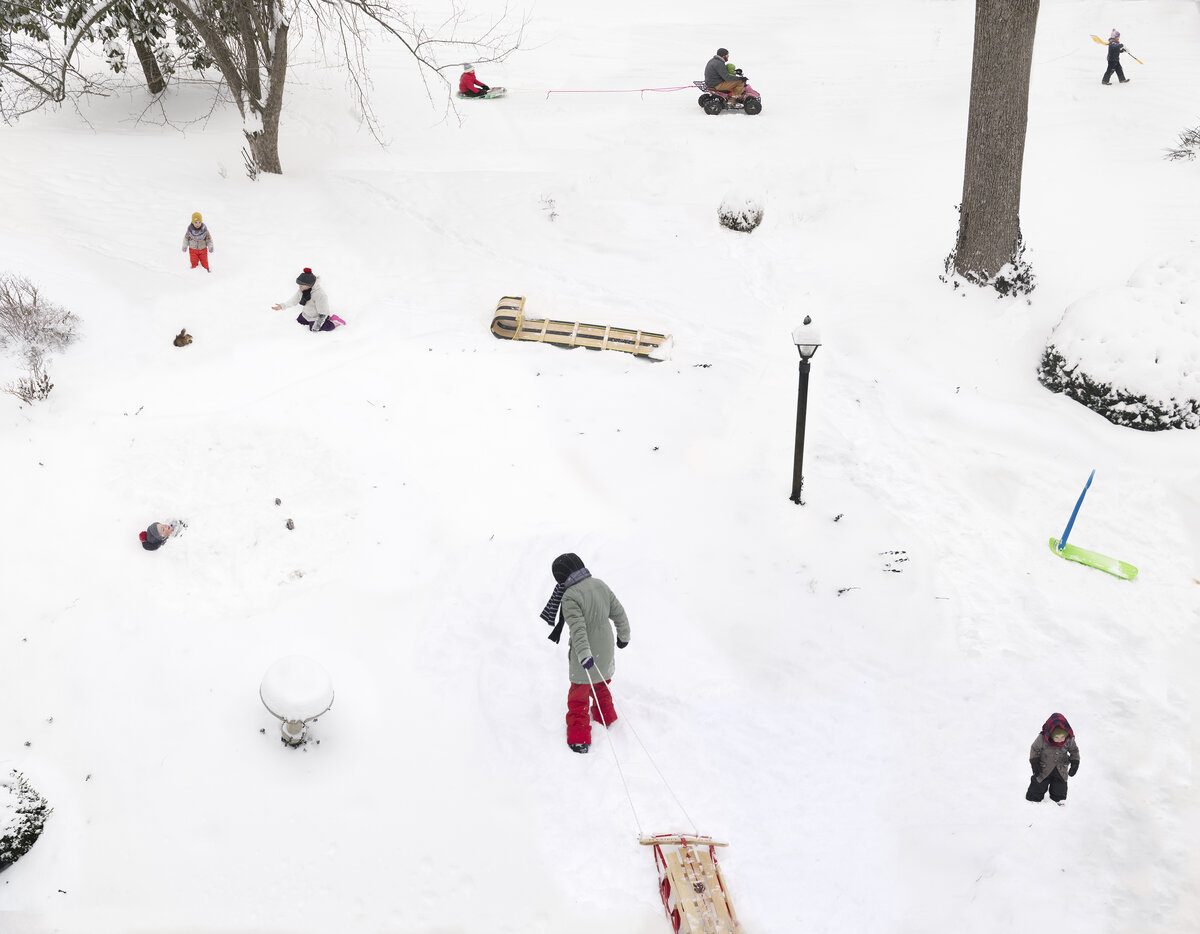 Aerial view of a snowy hillside dotted with people engaged in winter activities—sledding, playing, and walking—scattered across the white landscape with bare trees, a lamp post, and various sleds and toys visible from above.