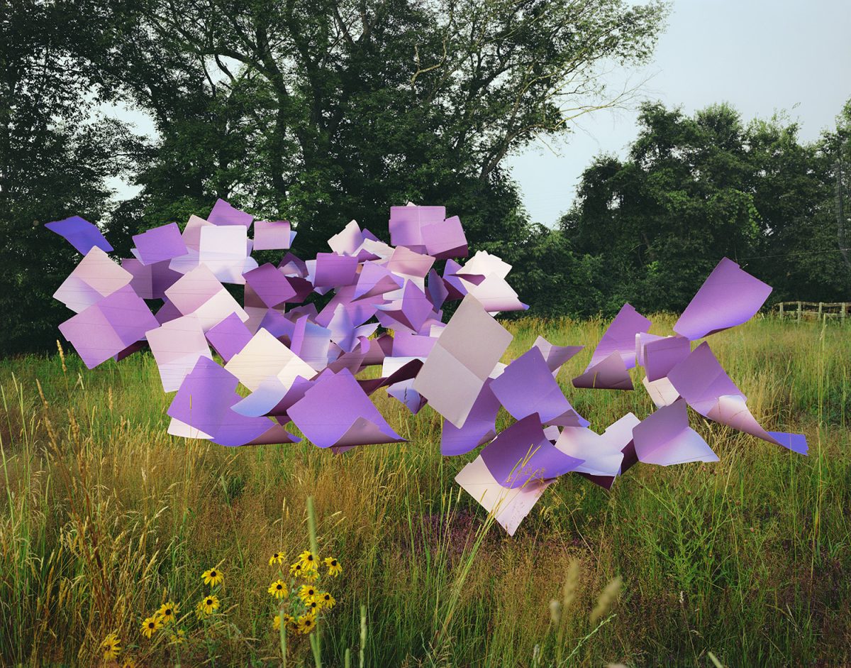 Large-scale sculptural arrangement of purple and white file folders positioned in a grassy meadow with wildflowers and trees in the background, creating an abstract three-dimensional composition that contrasts geometric forms with natural landscape.