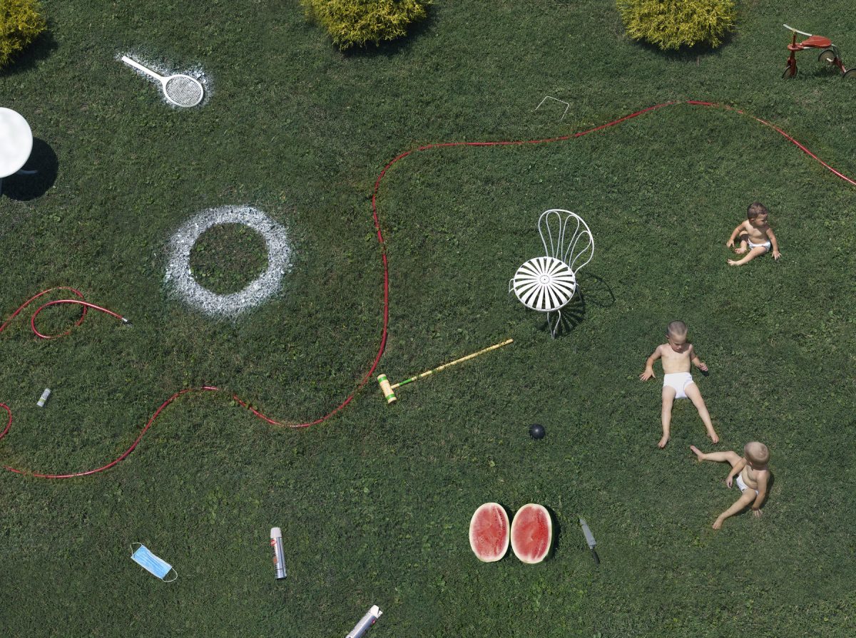 Aerial view of a lush green lawn scattered with everyday objects and three shirtless children at play. White chalk circles, a red garden hose, lawn furniture, a watermelon cut in half, gardening tools, and household items are arranged across the grass in a carefully composed domestic scene.