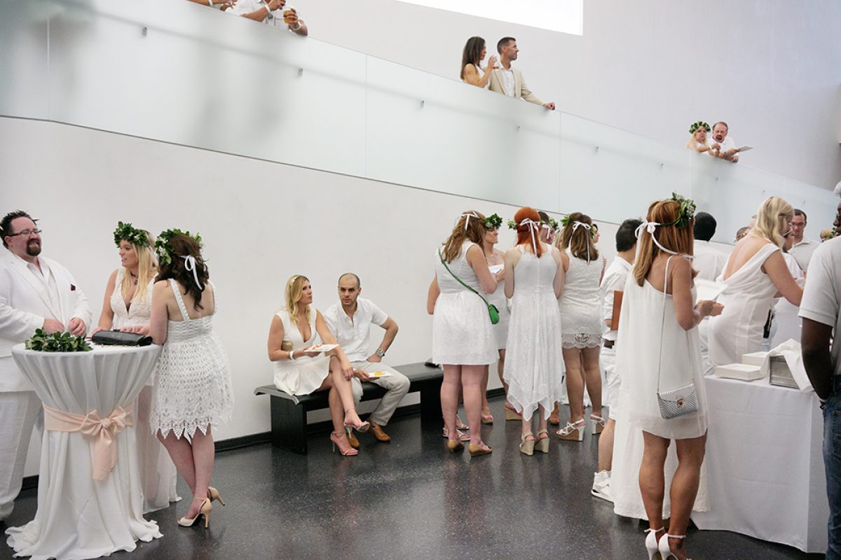 A large group of people dressed in white clothing gather in a modernist interior space with white walls and a dark floor. Guests mingle and socialize across multiple levels, with some standing on an elevated platform visible in the background, while others interact in the main hall below. Green floral garlands and white-draped cocktail tables accent the scene.