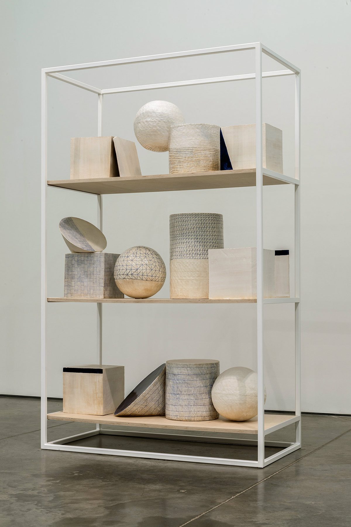 White steel display case with four wooden shelves containing an arrangement of geometric sculptural objects in natural materials including woven baskets, spheres, cylinders, and rectangular forms in cream, beige, gray, and black tones.