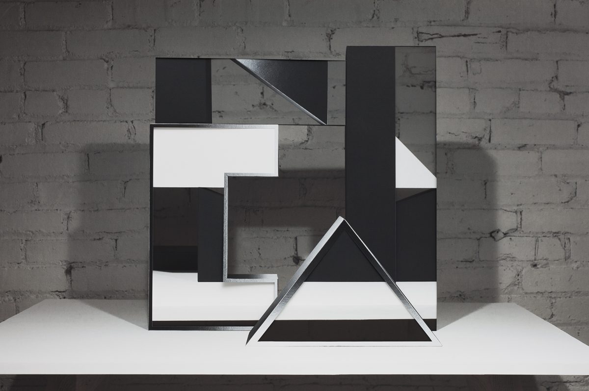 Abstract geometric composition of layered black and white paper forms arranged on a white platform against an exposed brick wall. Interlocking rectangles and a triangular shape create a three-dimensional collage with sharp angles and negative space, emphasizing contrast between dark and light surfaces.