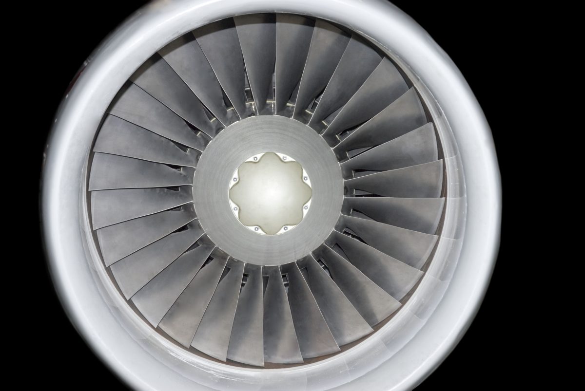 Front-facing view of a jet engine turbine with concentric metal blades radiating from a central hub, rendered in grayscale tones against a black background. The precise, symmetrical composition emphasizes the industrial geometry and reflective surfaces of the machinery.
