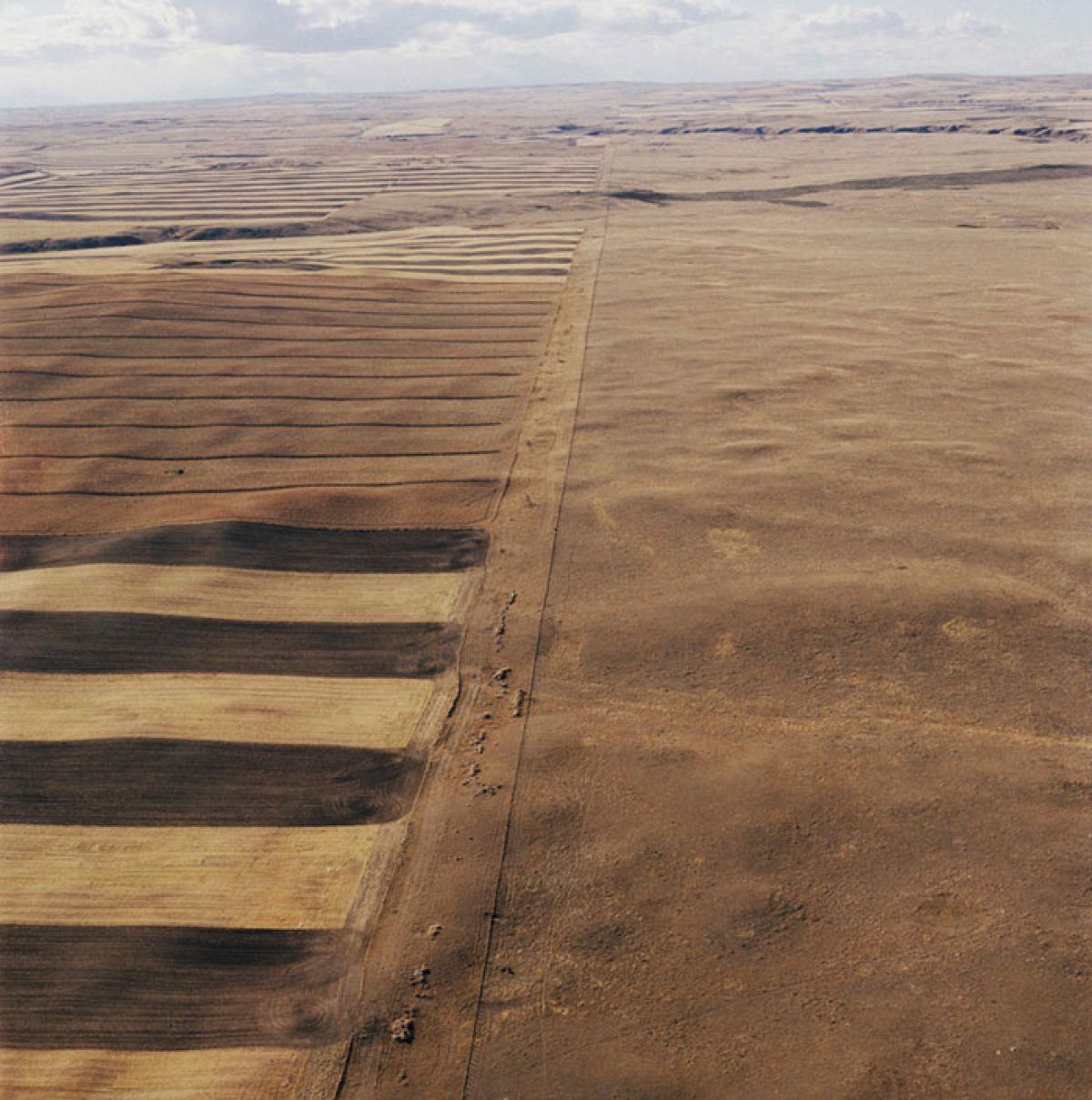 Aerial view of agricultural land divided by a straight dirt road, with plowed fields in varying shades of brown and tan on the left and an open, fallow field on the right. The flat, expansive landscape stretches toward the horizon beneath a cloudy sky.