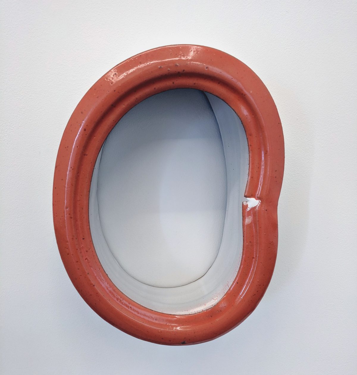 A terracotta-red ceramic form with an oval opening, featuring a smooth inner surface in contrasting gray-blue. The outer frame has a rounded, sculptural quality with visible surface texture and a small white chip on the right edge.