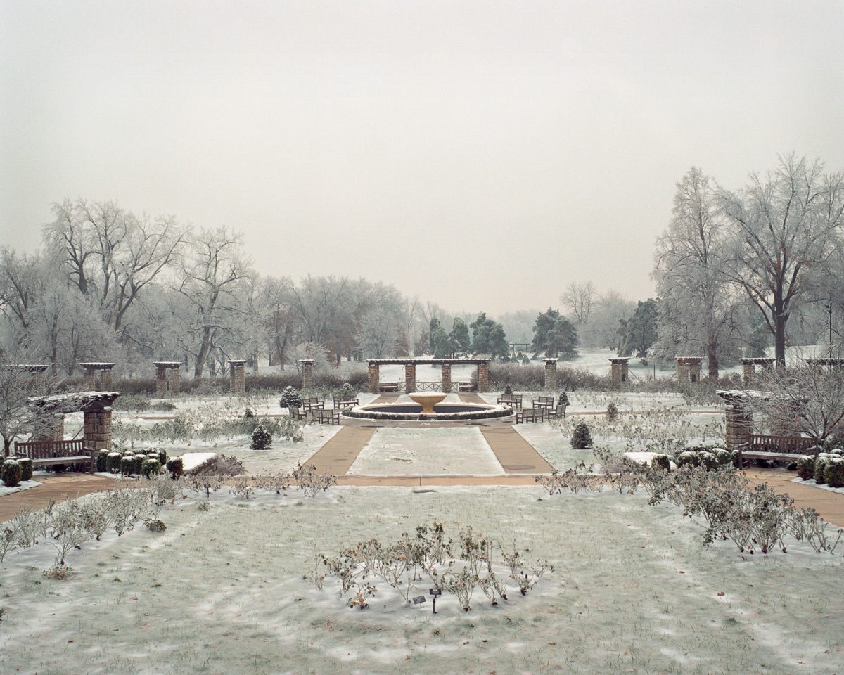 A frost-covered formal garden with a central circular fountain viewed down a symmetrical pathway. Bare trees frame the scene on both sides while brick structures and evergreens line the composition, all blanketed in ice and snow beneath an overcast sky.