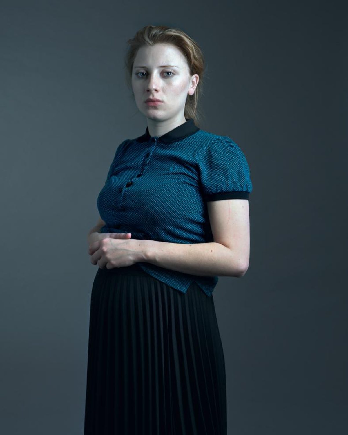 A woman with reddish-blonde hair stands against a neutral gray background, wearing a teal textured short-sleeved top with a black collar and a black pleated skirt. She gazes directly at the camera with a composed, contemplative expression, her arms crossed in front of her body.