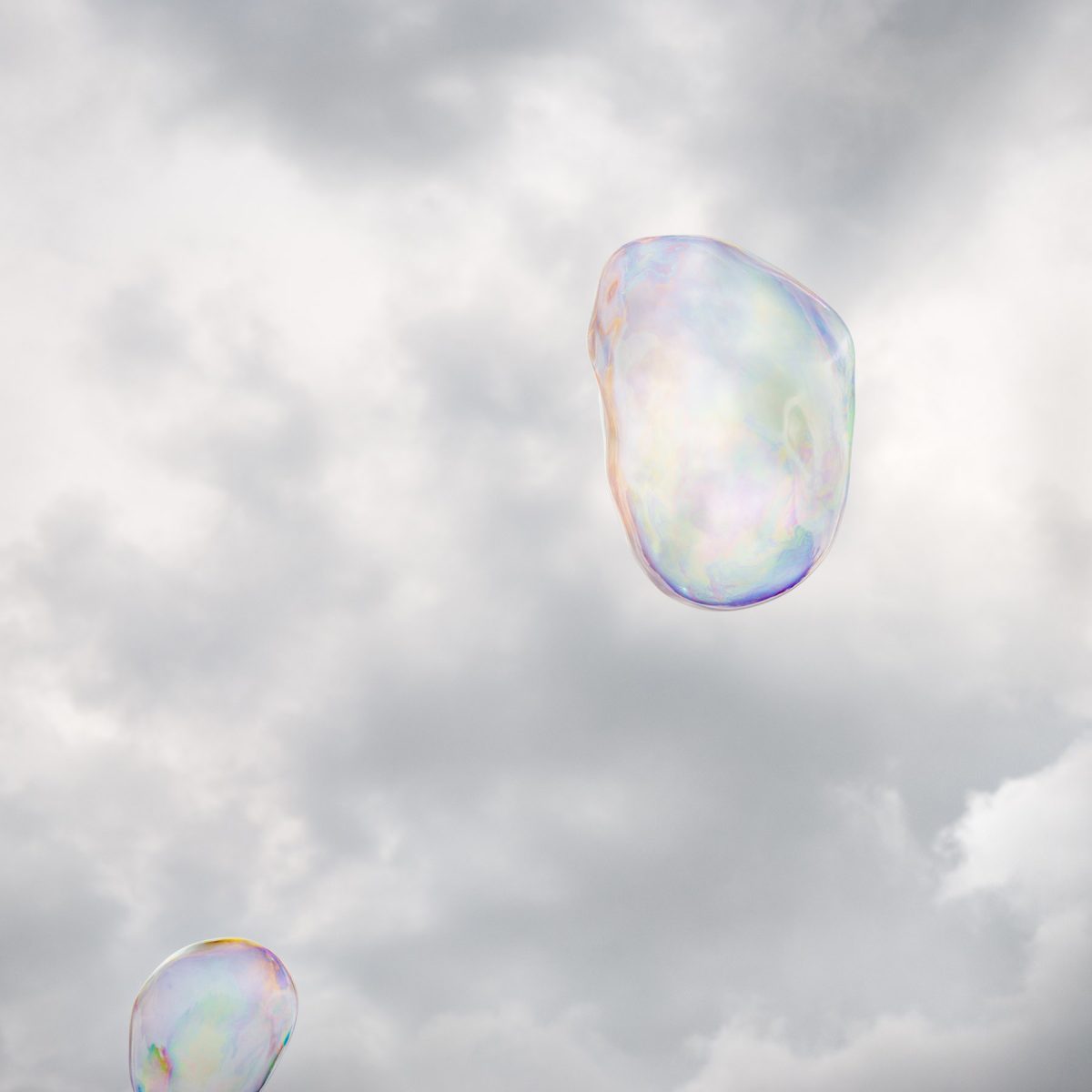 Two iridescent soap bubbles floating against a cloudy gray sky, their translucent surfaces reflecting soft pastel hues of blue, pink, yellow, and green.