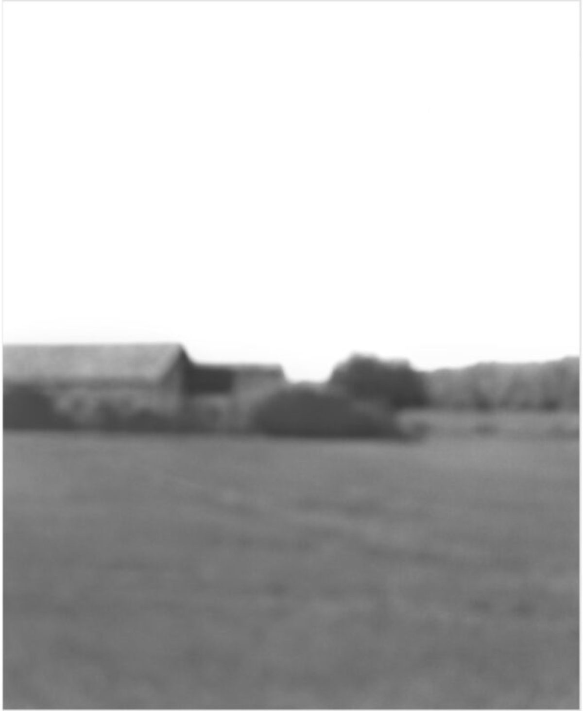 Monochromatic landscape photograph featuring a distant rural scene with farm buildings and trees along the horizon, rendered in soft focus with a hazy, atmospheric quality. The composition is dominated by an empty foreground of flat terrain beneath a pale sky.