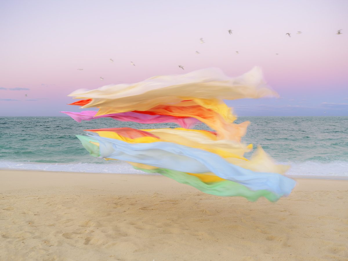 Billowing sheets of colorful silk—layered in shades of white, cream, pink, red, orange, yellow, blue, and green—dance in the wind on a sandy beach at dusk, with the ocean and a purple-pink sky visible beyond.