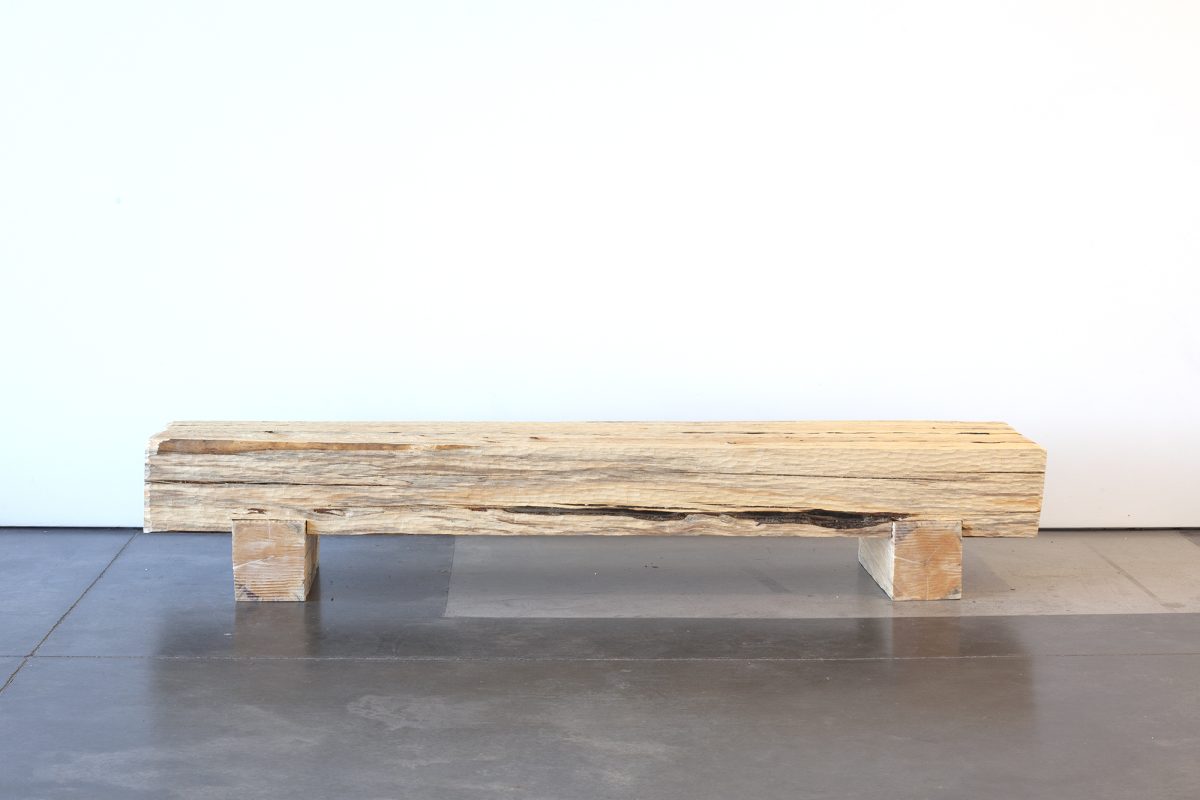 A long wooden bench made from a single weathered log with a dark burn scar running across its top surface, supported by two rectangular wooden blocks, sits on a polished concrete floor against a white wall.