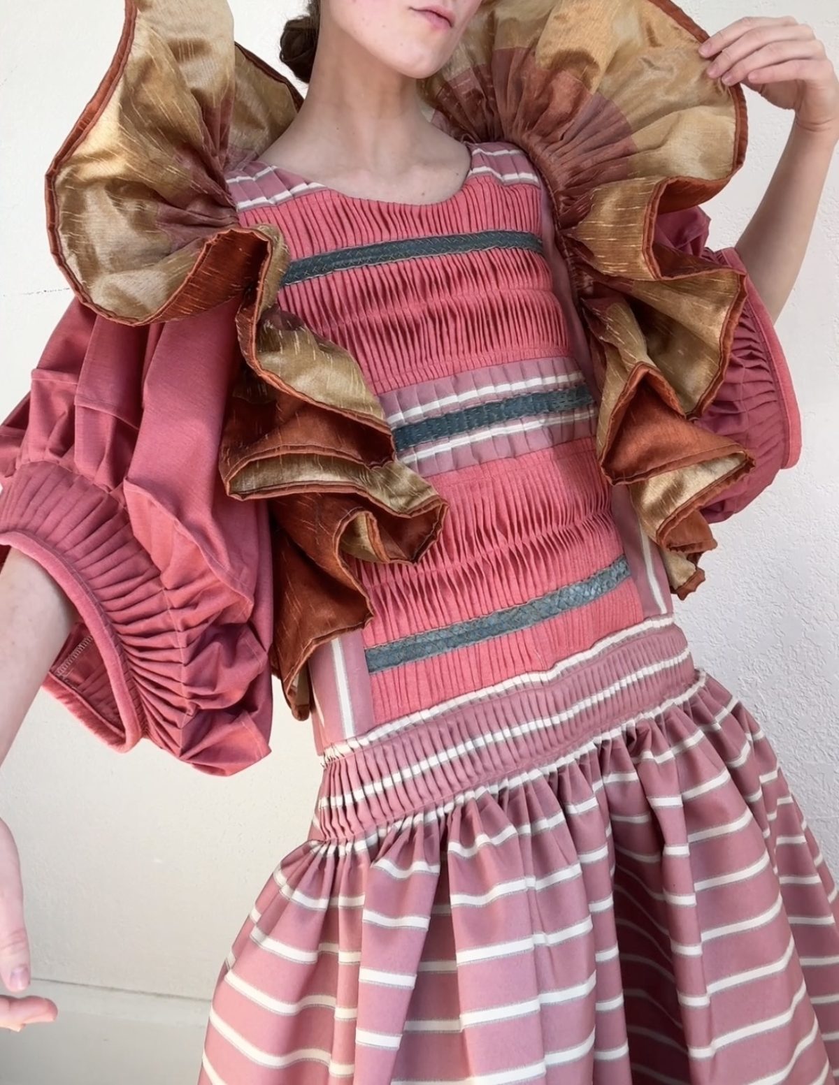 A figure wearing a voluminous pink dress with horizontal striped pleating and white banding stands with arms raised, displaying dramatic ruffled shoulders in contrasting gold and burgundy taffeta. The composition showcases intricate textile craftsmanship through hand-pieced pleats and embroidered details throughout the garment.