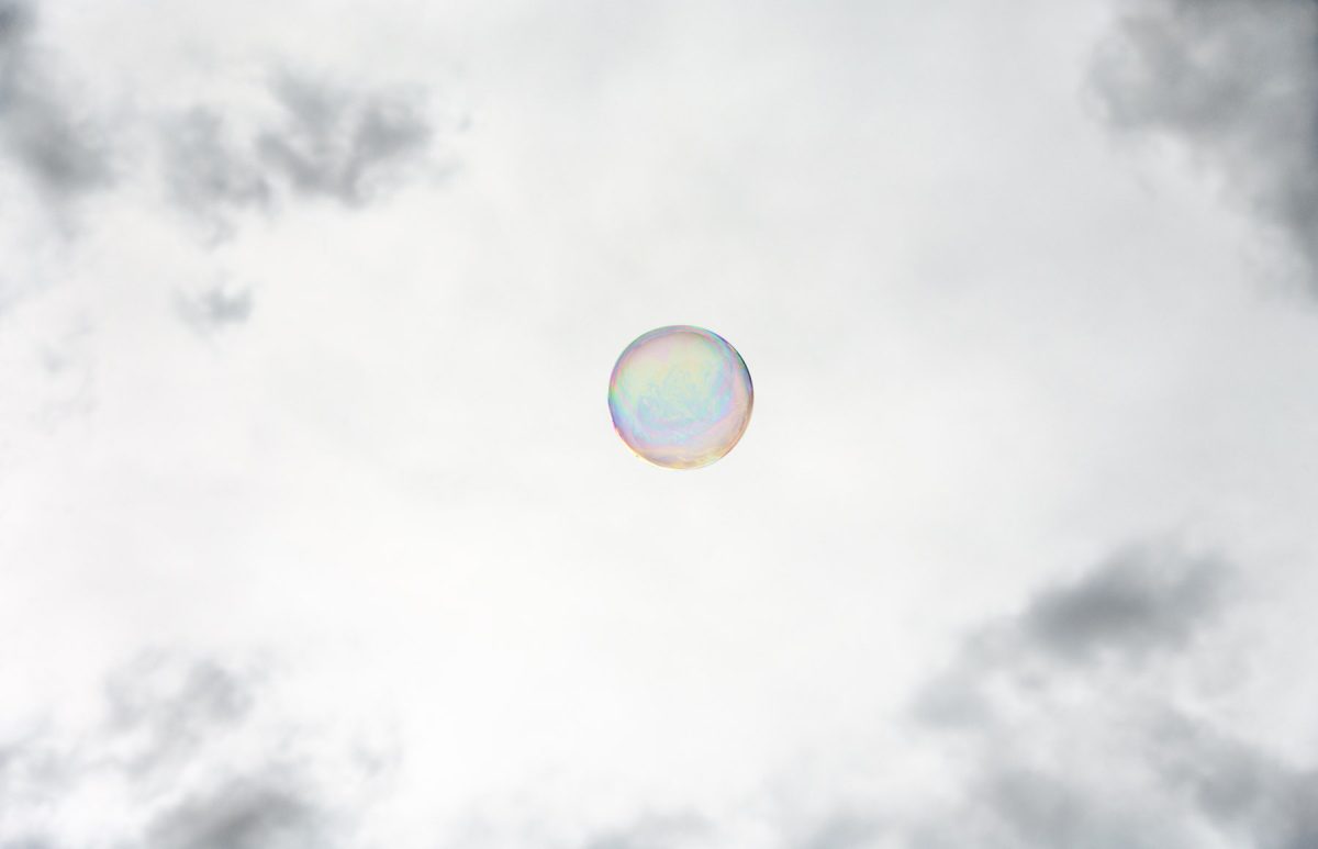 A single iridescent soap bubble floats against a soft, overcast sky with diffuse clouds visible at the edges. The bubble's translucent surface displays subtle rainbow hues of blue, pink, and yellow in its curved form.
