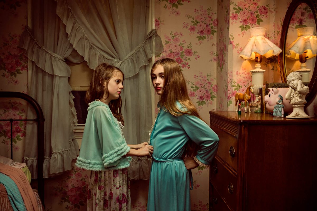 Two young girls in turquoise robes stand facing each other in a vintage bedroom decorated with rose-patterned wallpaper and sheer curtains. Warm lamplight illuminates classical decorative objects on a wooden dresser behind them, creating a nostalgic, staged tableau.