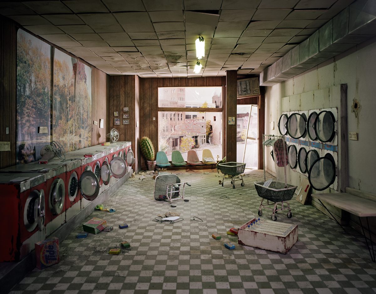 An abandoned laundromat interior with red and black washing machines lining the left wall and dryer units mounted on the right. The checkered floor is scattered with toys and debris, while colorful plastic chairs and laundry baskets sit near large windows overlooking an urban cityscape. Fluorescent ceiling lights illuminate the deteriorating space with peeling paint on pale green walls.