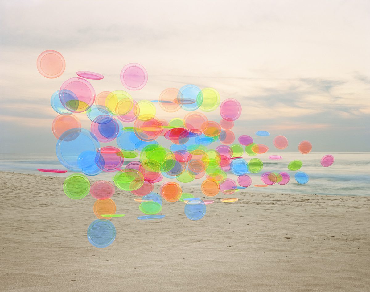 Translucent colored plastic discs and utensils in vibrant hues of blue, pink, green, yellow, and orange are arranged on a sandy beach, creating an abstract composition against a calm ocean and overcast sky.