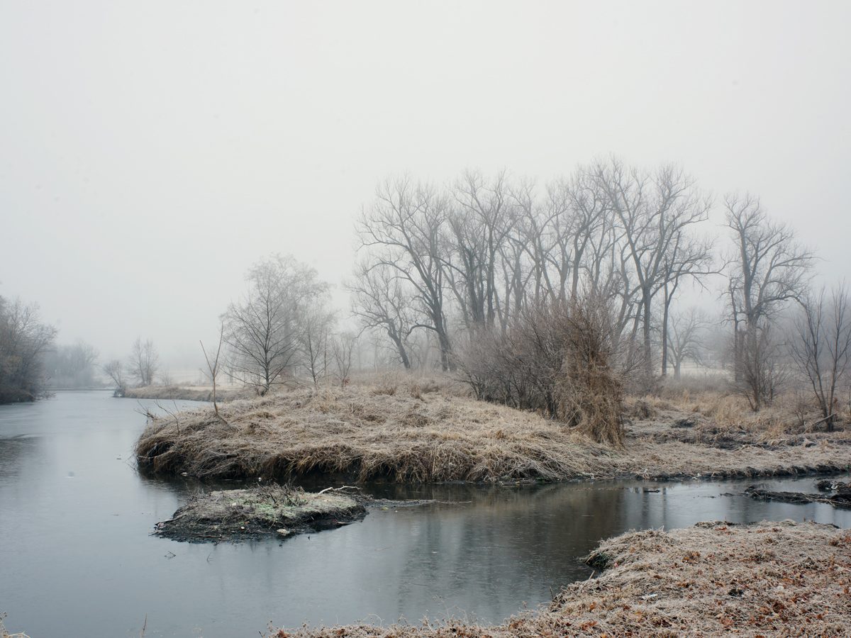 A misty riverside landscape with bare, leafless trees receding into fog. The composition features a shallow creek or waterway in the foreground with sandy banks and rocky outcrops, bordered by dormant vegetation and a dense stand of skeletal trees along the far bank. The overall palette is muted in grays, browns, and pale tones, creating a somber, winter atmosphere.
