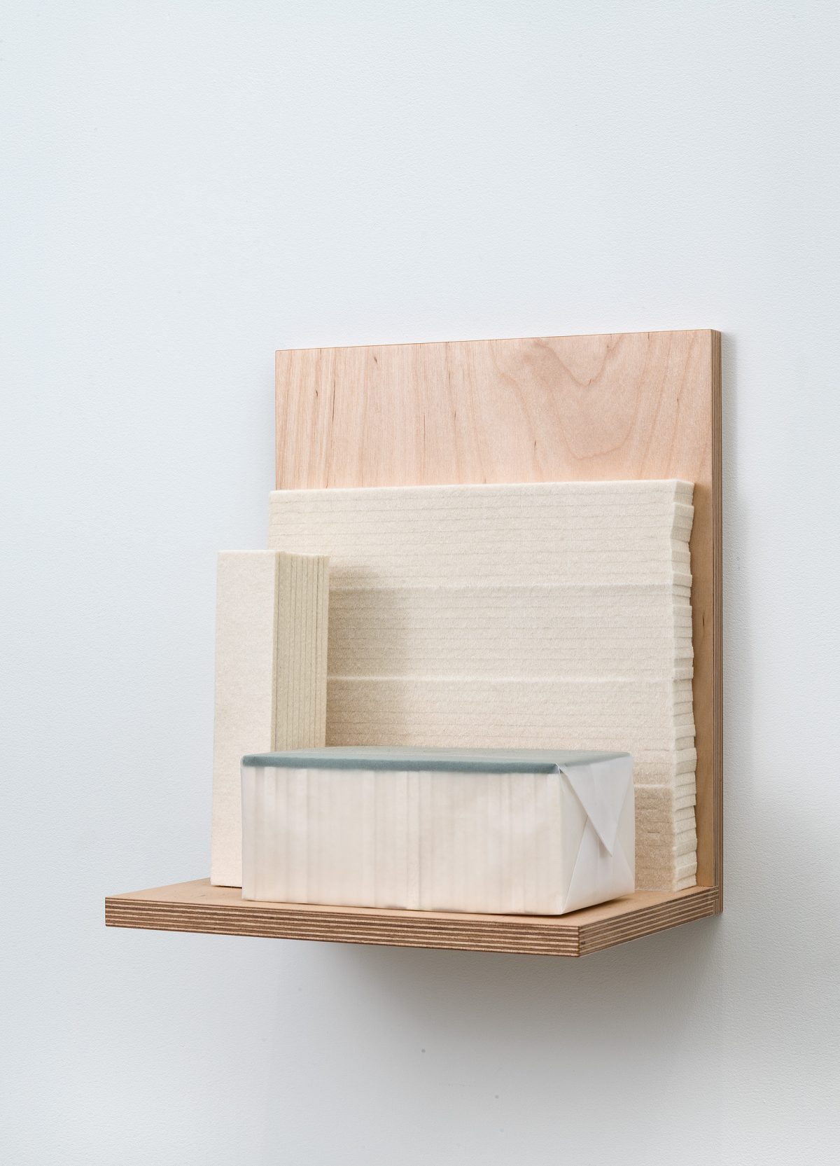 A wooden wall-mounted shelf unit constructed from light plywood and natural wood veneer, featuring geometric compartments and a cream-colored fabric-wrapped box with a blue-gray felt edge resting on the lower shelf.