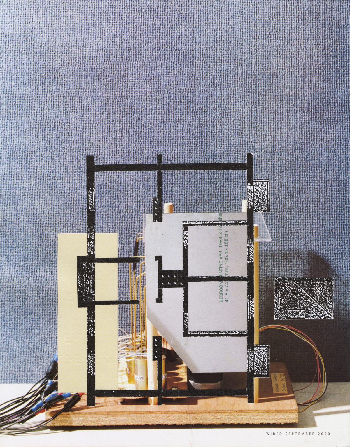 A three-dimensional collage construction mounted on a wooden base features black linear frameworks resembling architectural models, cream-colored paper planes, white rectangular forms with printed text, and wired electronic components in orange and red. Against a textured blue-gray backdrop, the assembled elements create a layered, abstract composition blending architectural, industrial, and mixed-media aesthetics.