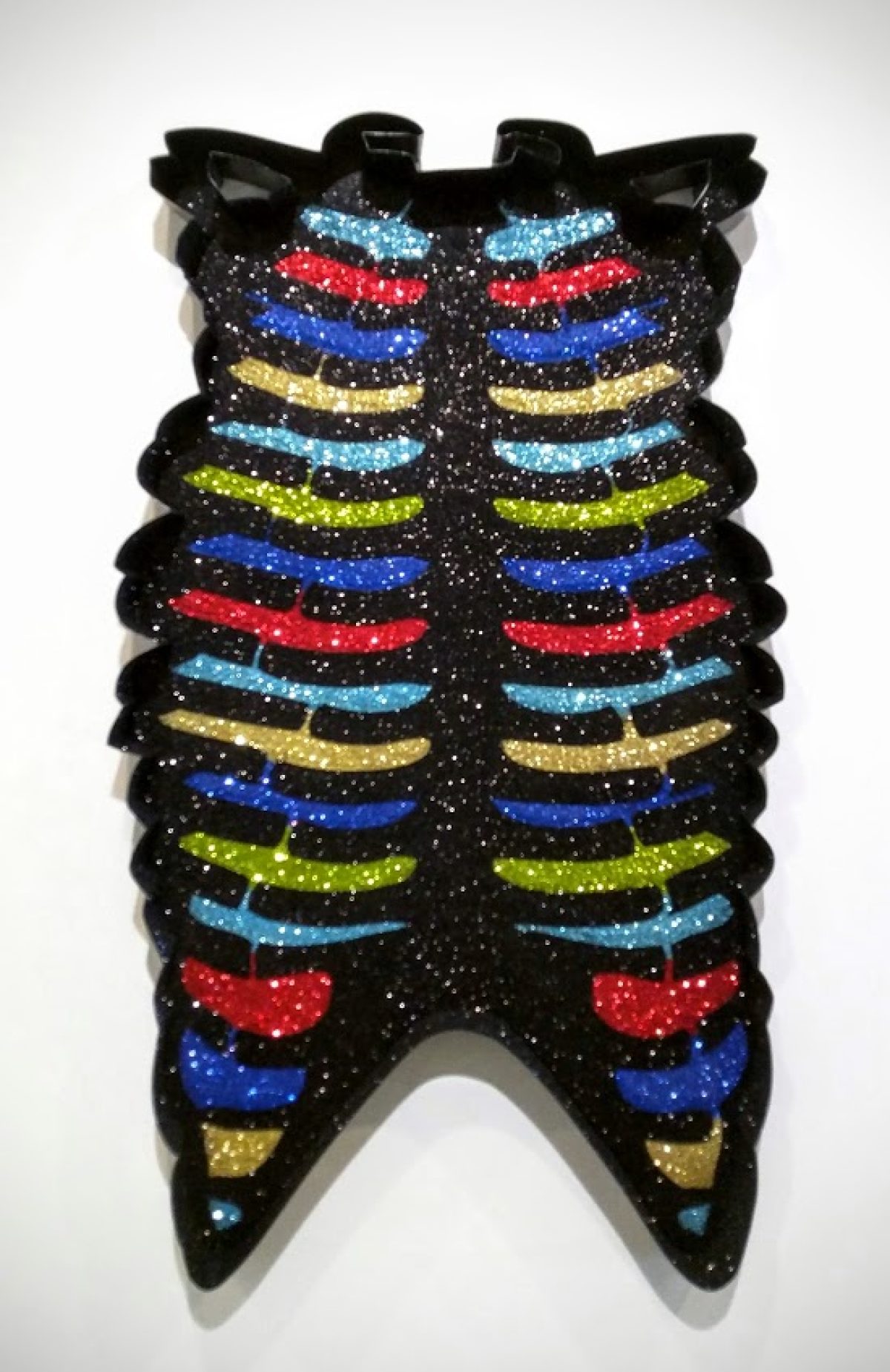 A skeletal ribcage rendered in black with symmetrical horizontal bands of glitter in vibrant colors—cyan, red, blue, gold, and chartreuse—suspended in glossy resin. The form is frontal and vertical with pointed ends at top and bottom, mounted against a white wall.