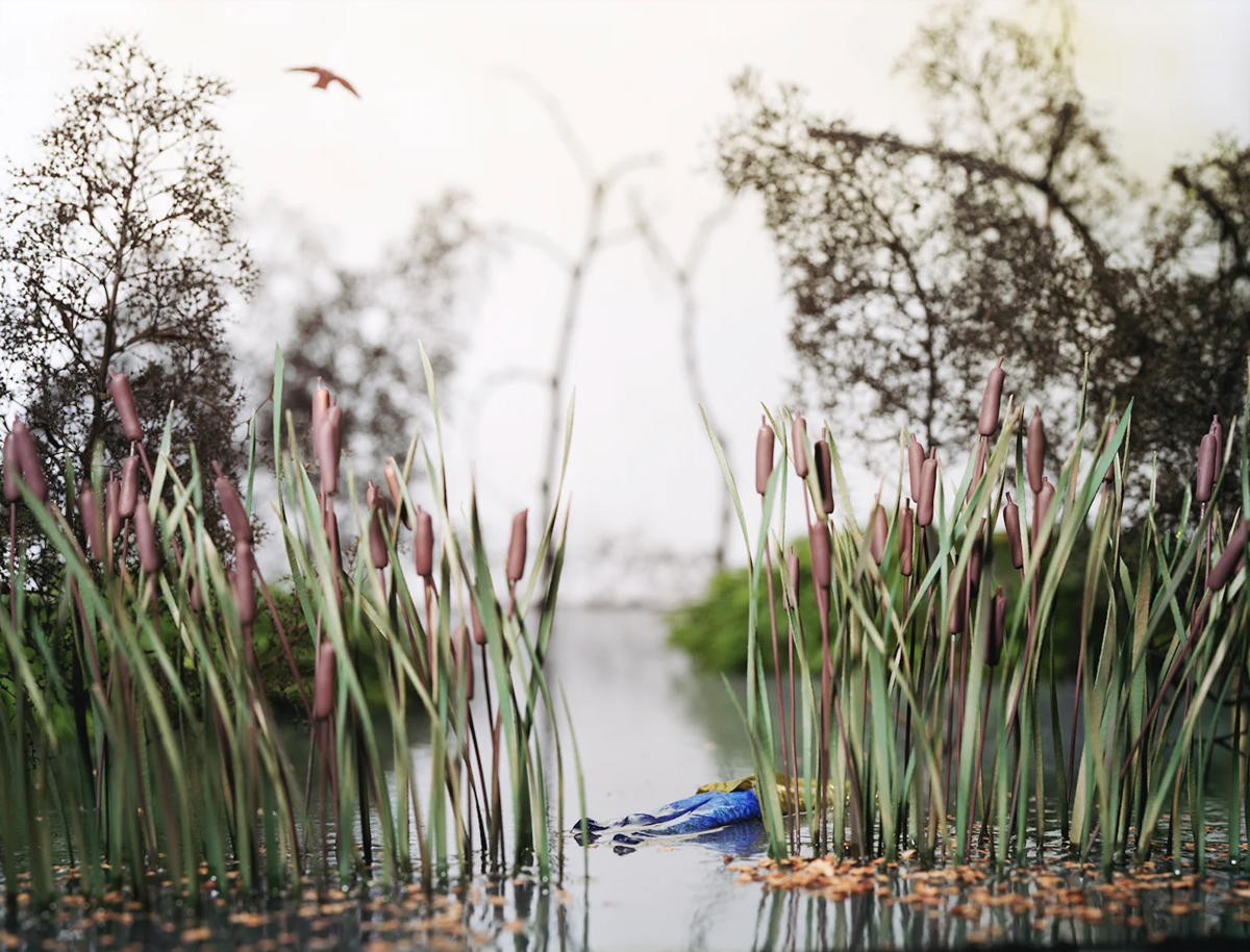 A wetland scene with purple-budded cattails in sharp focus framing either side of a shallow waterway. In the background, flowering trees with delicate white blossoms blur softly, while a blue crocodile or alligator floats partially submerged in the water between the reeds. A bird flies in the overcast sky above.