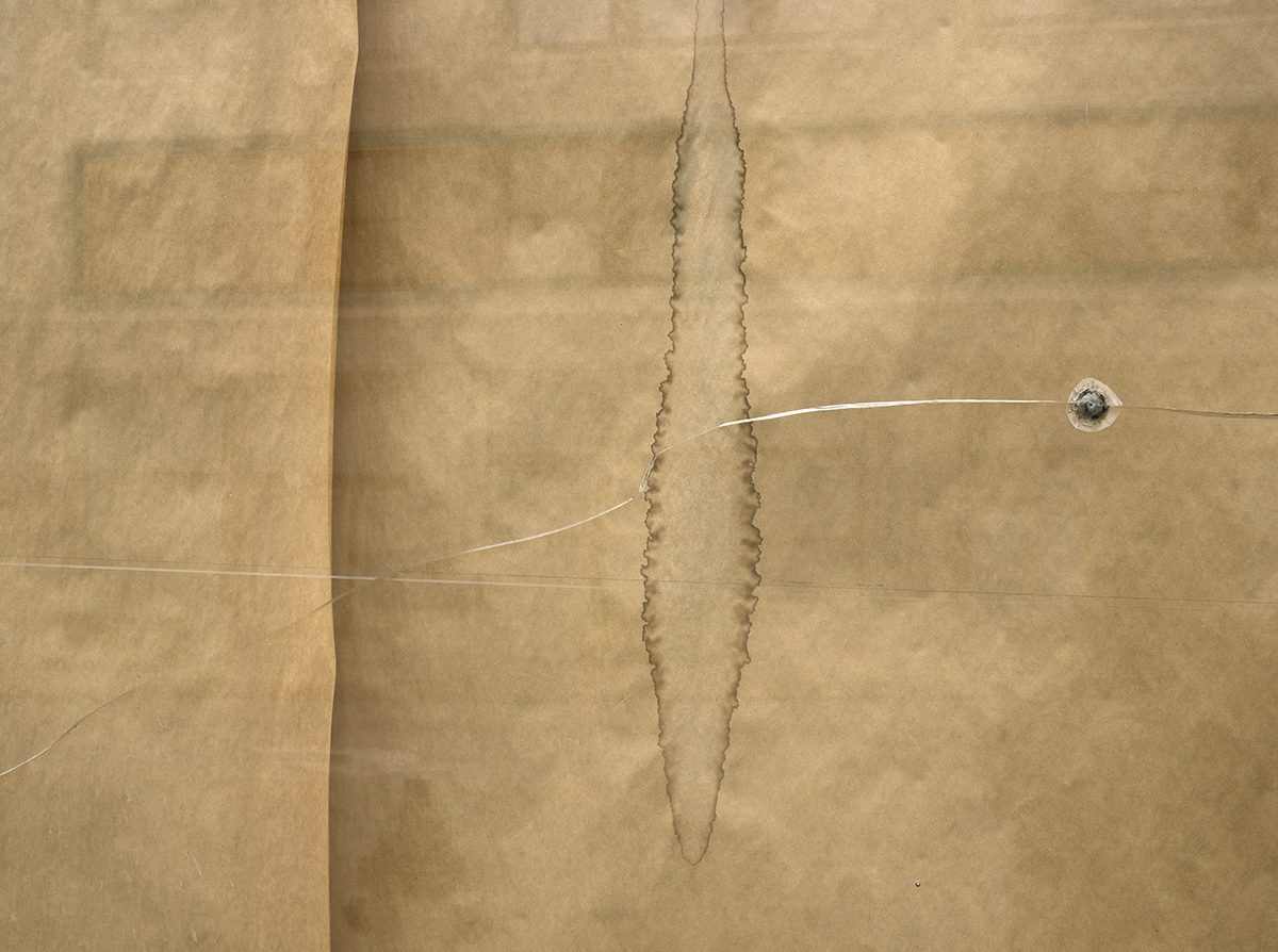 Folded tan paper with a vertical torn seam down the center, accompanied by two thin translucent lines extending across the composition, each terminating at a small spherical form on the right side.