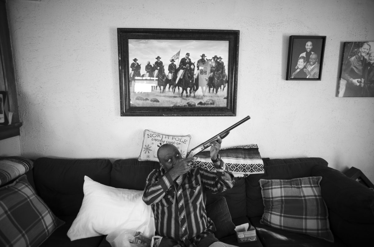 Black and white photograph of a person seated on a dark couch holding a rifle, positioned beneath framed photographs and artwork on a white wall. The composition captures an intimate domestic interior with patterned pillows and blankets, while the subject's pose creates a stark juxtaposition between the domestic setting and the weapon.
