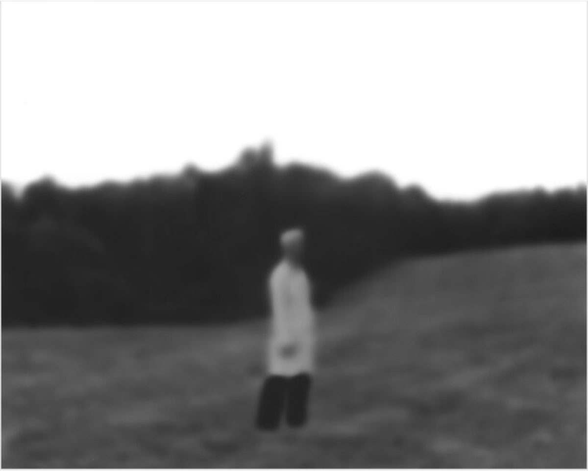 A solitary figure in a white shirt and dark pants stands in a vast, empty field, facing away from the camera toward a dark treeline on the horizon. The black and white photograph has a soft, slightly out-of-focus quality that emphasizes the isolation of the lone person in the expansive landscape.