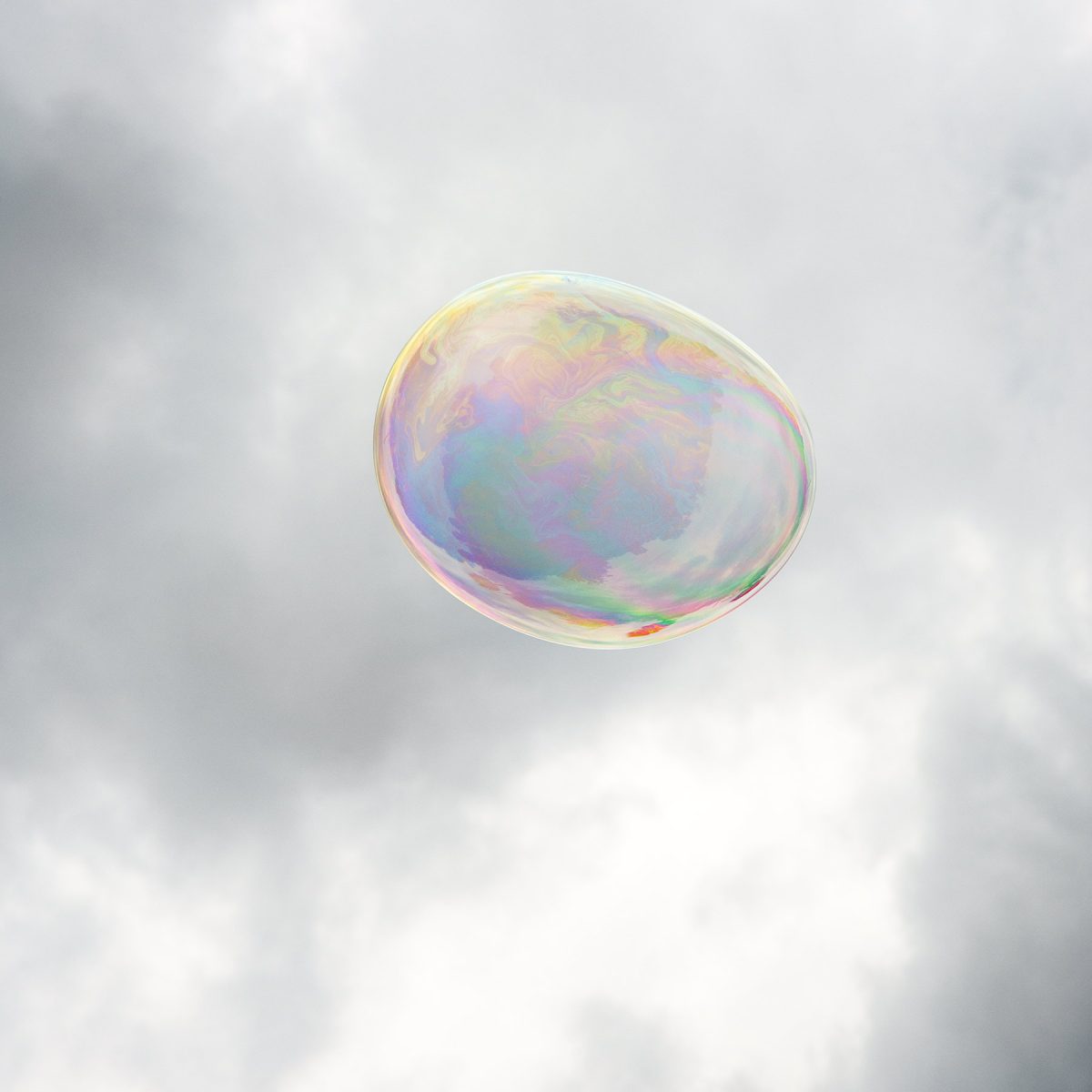A translucent soap bubble floats against a soft, cloudy sky, its spherical surface shimmering with iridescent bands of pink, purple, blue, and gold that shift across its curved form.