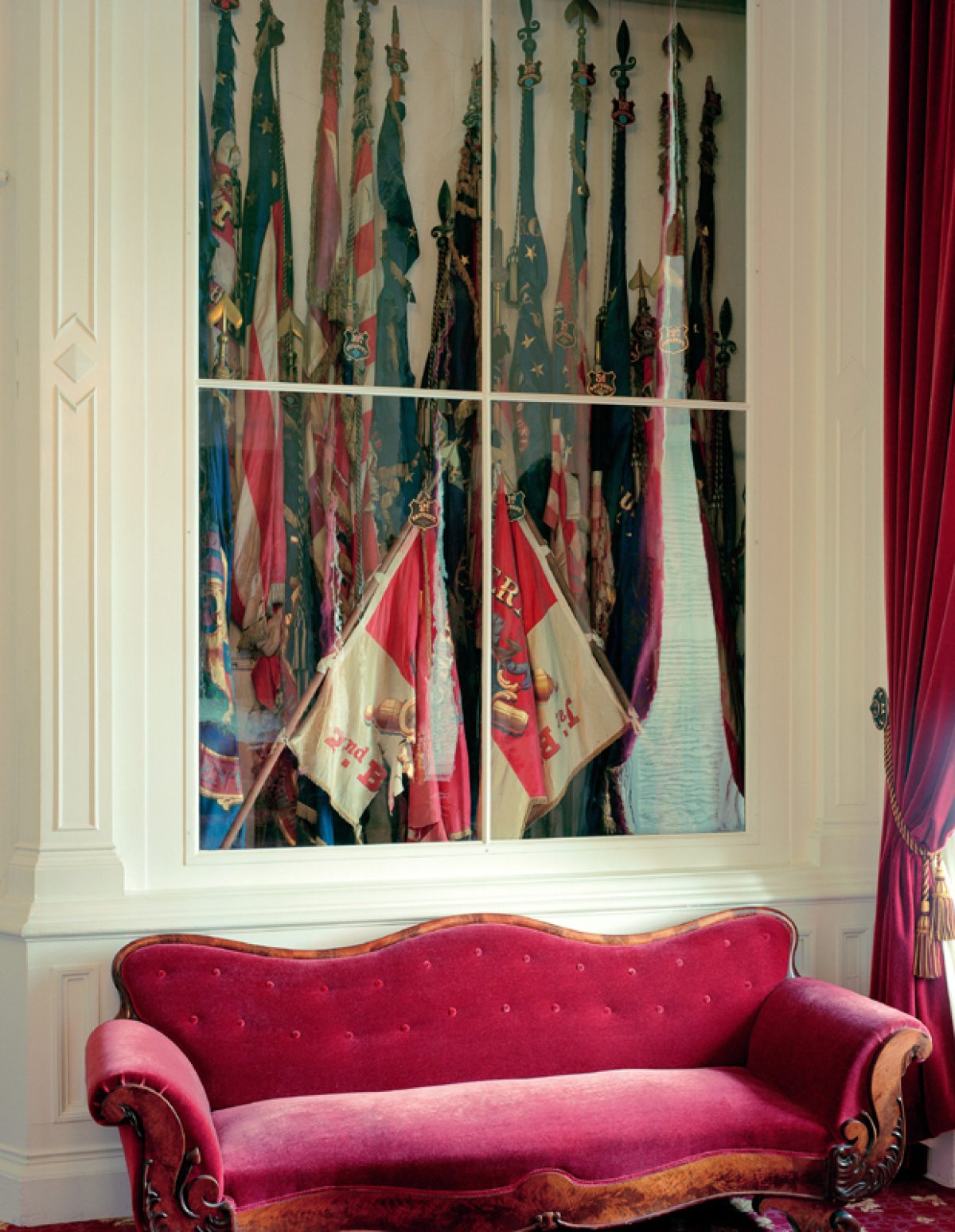 A framed photographic print depicting a collection of ornate flags and banners in jewel tones—greens, reds, and blues—displayed vertically. Below the print sits a luxurious magenta velvet settee with curved arms and decorative nail head detailing, positioned against a cream-colored wall with red velvet curtains visible to the right.