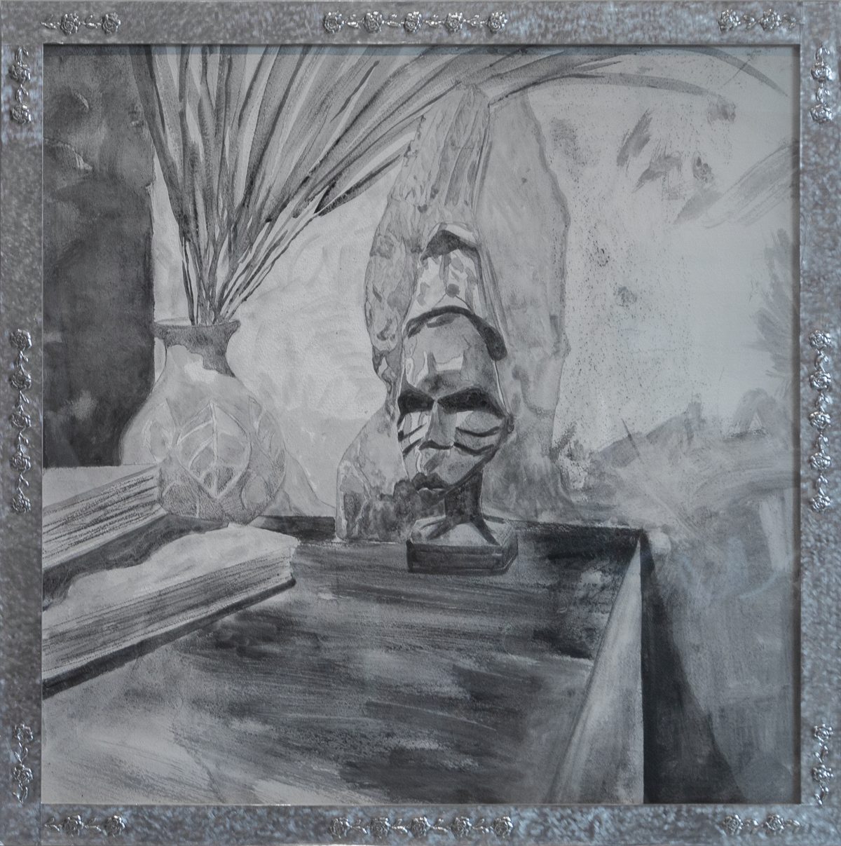 A graphite drawing on gesso depicting a sculptural bust with pronounced facial features and a contemplative expression, positioned on a wooden surface. Dried palm fronds in a decorative vessel are visible to the left, while atmospheric marks and gestural strokes create depth in the background. The work is presented in an ornate silvered frame.