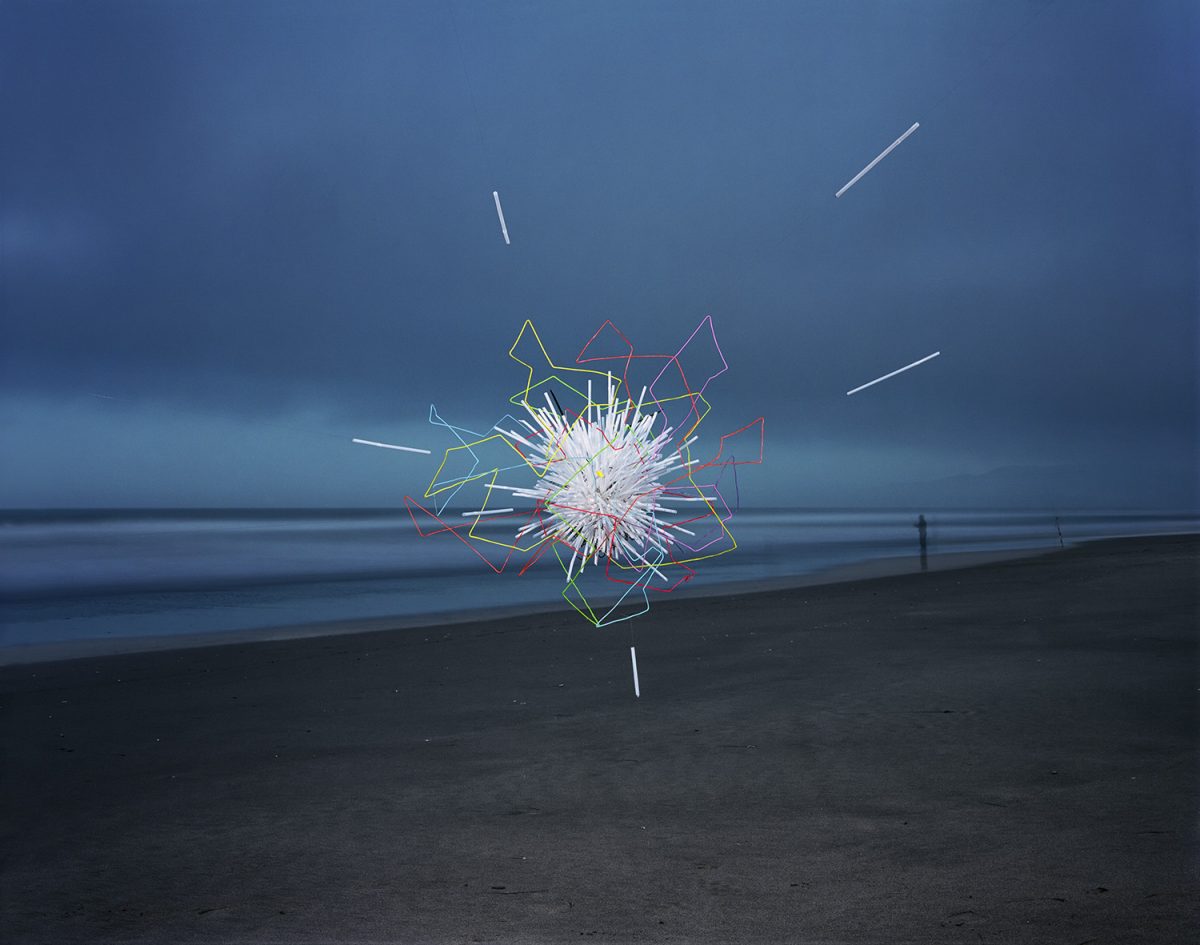 Neon-outlined geometric shapes in yellow, red, pink, and white radiate outward from a bright central burst on a dark beach, with horizontal straws extending across the composition against a moody blue-grey ocean and sky.