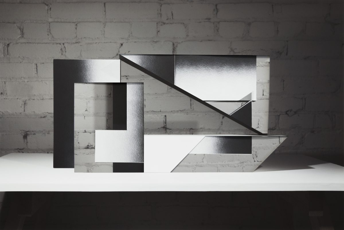 An abstract sculptural composition of layered black and white rectangular forms arranged on a white platform against a white brick wall. The overlapping planes create sharp angles and diagonal lines, with contrasting matte and reflective surfaces that cast distinct shadows.