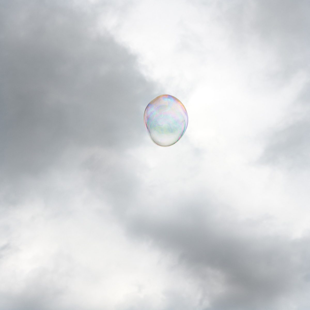 A single iridescent soap bubble floats against a cloudy gray and white sky, its translucent surface reflecting soft pastel hues of pink, blue, and green.