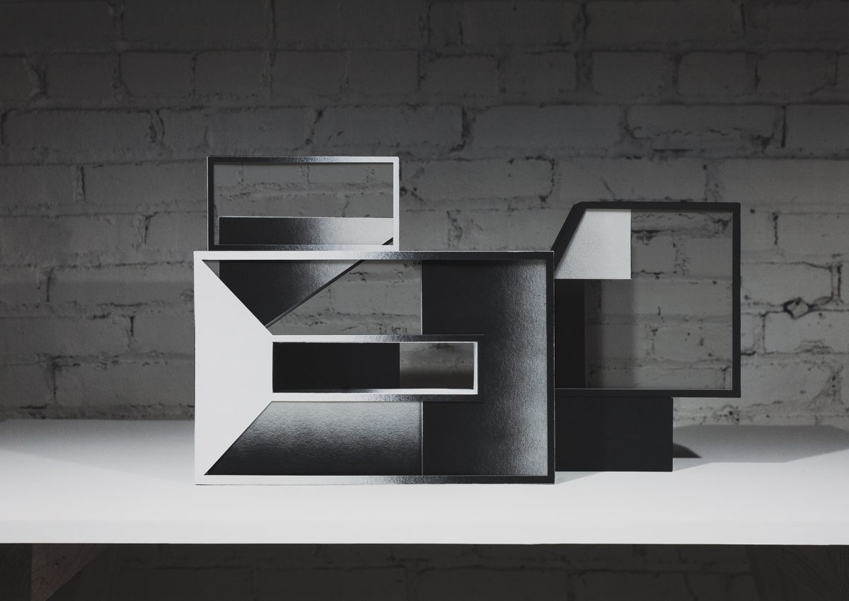 Abstract geometric composition featuring interlocking rectangular frames in black and white, layered to create a three-dimensional effect against a weathered brick wall background. The fragmented, overlapping planes suggest architectural forms with varying depths and contrasting tones.