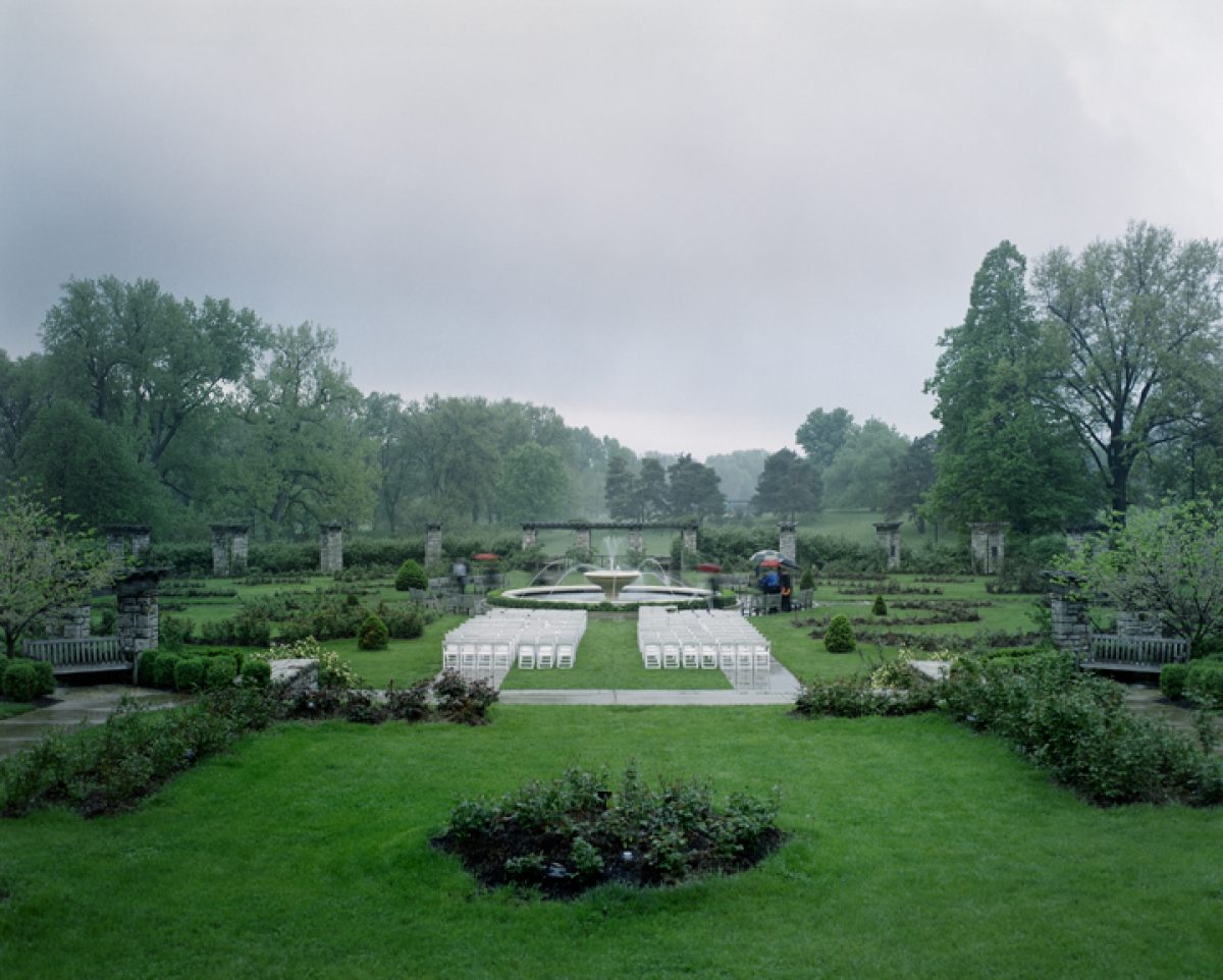 A formal garden ceremony setup with white chairs arranged in rows beneath a white tent pavilion, flanked by manicured hedges and classical stone columns. Lush green lawn and mature trees frame the scene under an overcast sky, with water visible in the distance.