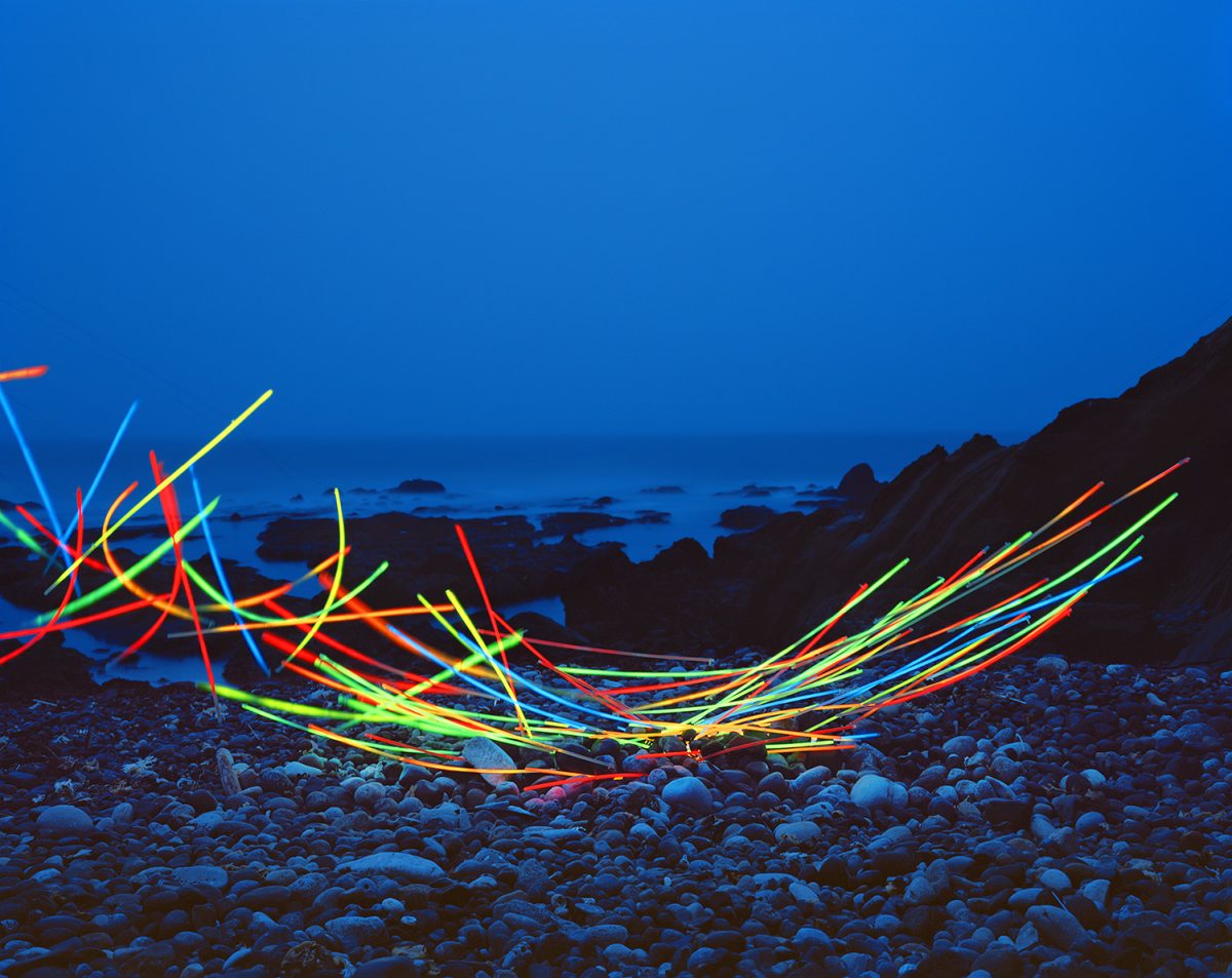 Neon glow sticks arranged in tangled, radiating patterns across a rocky beach at dusk, their bright red, yellow, green, and blue lines creating dynamic linear forms against the darkened landscape and twilight sky.