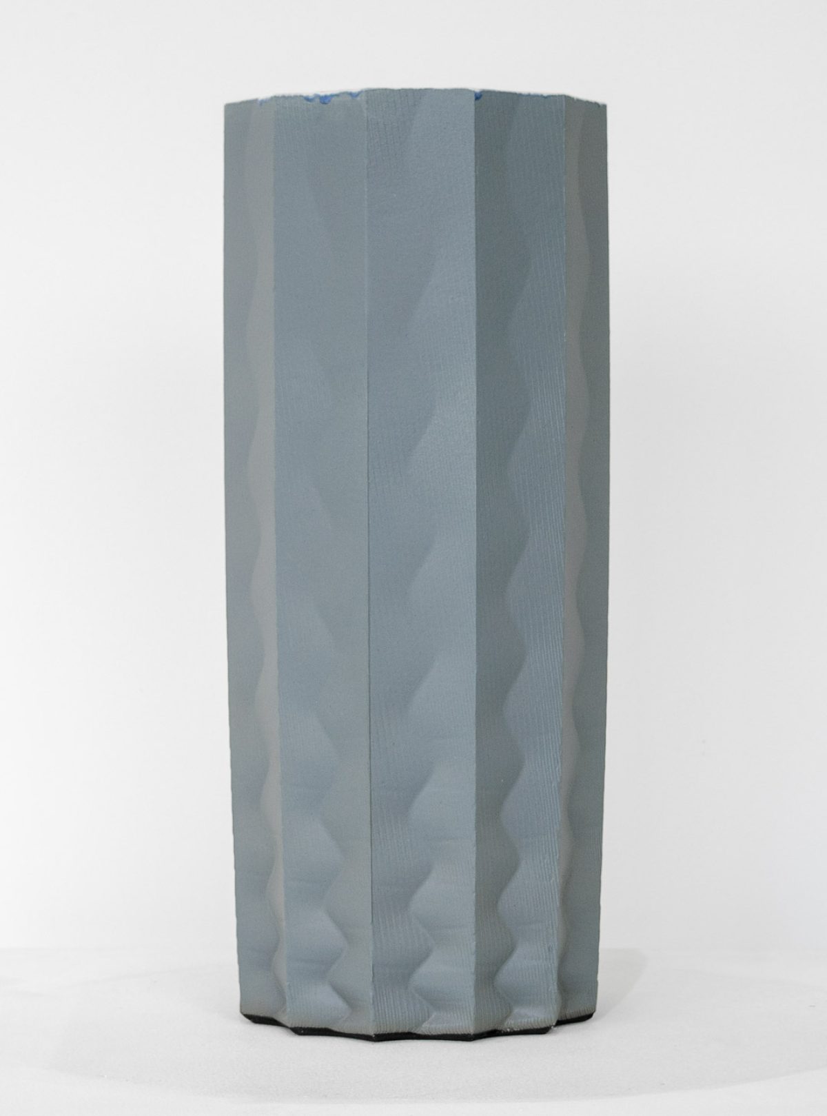A tall cylindrical porcelain form with vertical ridged panels in soft gray and white tones, featuring subtle wave-like undulations across its surface. The sculpture stands against a neutral background, its organic, fluted structure creating areas of shadow and depth.